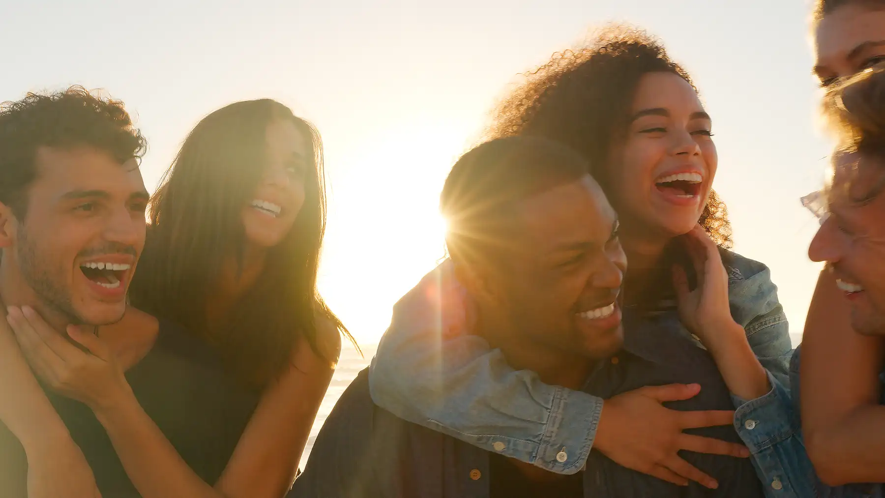 A diverse group of friends laughing together, arms around each other, backlit by warm evening light